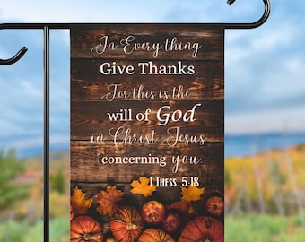 May include: A decorative garden flag with a wooden plank background and the text "In Everything Give Thanks". The flag features a fall theme with pumpkins and leaves at the bottom. The flag is hanging on a black metal stand.