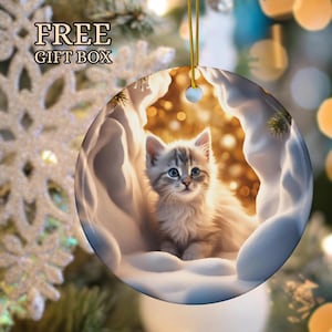 May include: A white ceramic Christmas ornament with a cute kitten sitting in a snowy cave. The kitten has blue eyes and is looking at the camera. The ornament is decorated with a gold string and a gold snowflake.