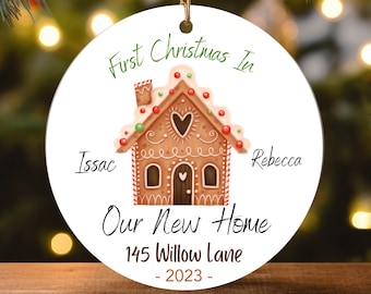 May include: A white round Christmas bauble featuring a gingerbread house illustration. The text reads "First Christmas In Our New Home 145 Willow Lane - 2023 -" with the names "Issac" and "Rebecca" on either side of the house.