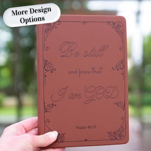 May include: A rectangular, rust-colored notebook with a dark brown engraved border and the text "Be still and know that I am GOD" followed by "Psalms 46:10" in the center. The cover features a smooth, matte finish with decorative corner accents.