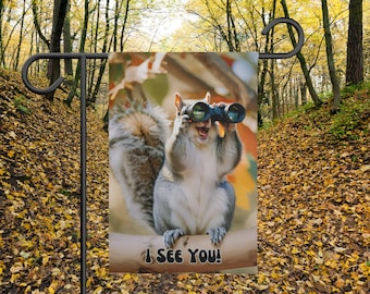 Könnte beinhalten: Eine Gartenflagge mit einem Eichhörnchen, das ein Fernglas trägt und den Text "Ich sehe dich!" Das Eichhörnchen sitzt auf einem Ast vor einem verschwommenen Hintergrund aus Herbstlaub.