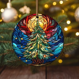 Puede incluir: Un adorno navide&ntilde;o redondo con un dise&ntilde;o de vitral de un &aacute;rbol de Navidad verde con un sol amarillo y un cielo azul. El adorno tiene el a&ntilde;o 2023 escrito en &eacute;l.