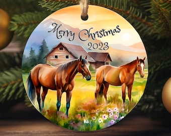 May include: A round Christmas bauble featuring a watercolour painting of two brown horses in a field with a barn in the background. The bauble has the text "Merry Christmas 2023" on it.