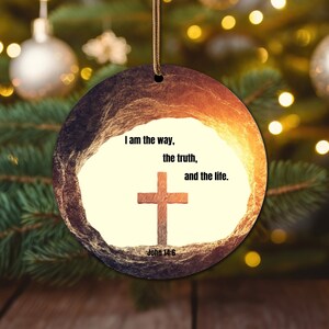May include: A round, wooden Christmas bauble with a rustic, textured surface. The bauble features a white background with a brown cross and the text "I am the way, the truth, and the life. John 14:6".