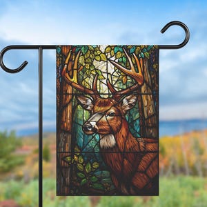May include: A garden flag displaying a stained-glass style image of a deer with large antlers. The deer is set against a backdrop of a forest with trees and foliage in shades of brown, green, and blue. The flag is mounted on a black metal stand.