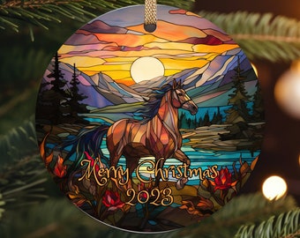 May include: Round Christmas ornament with a stained-glass design featuring a horse running near a lake, mountains, and a sunset. The ornament has the text "Merry Christmas 2023" in gold script and is hanging from a gold ribbon.