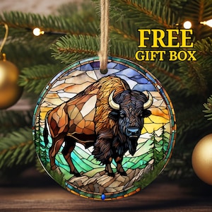 May include: A round, stained glass Christmas ornament featuring a brown and black bison standing on a rocky outcrop. The background is a colorful sunset with green trees.