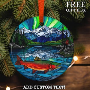 May include: Round Christmas ornament with a colorful stained-glass design featuring a lake scene with a red and green fish, mountains, and the Northern Lights. The ornament has an orange ribbon for hanging and the text "FREE GIFT BOX" and "ADD CUSTOM TEXT!"