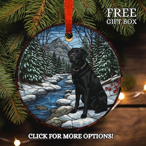 May include: Round Christmas ornament featuring a black Labrador Retriever sitting near a snowy stream. The ornament has a winter landscape scene with evergreen trees and a mountain in the background. The ornament is hanging from a red ribbon. Text on the ornament says "FREE GIFT BOX" and "CLICK FOR MORE OPTIONS!"