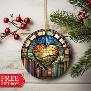 Op de afbeelding: Ronde kerstbal met een glas-in-loodontwerp met een hartvormig raam met een zonsondergangsscene en een rij boeken aan de onderkant. De kerstbal hangt aan een touwtje.