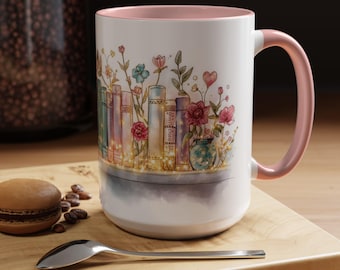 May include: White ceramic mug with a pink interior and handle. The mug features a watercolour design of books with flowers and fairy lights. A macaron, coffee beans, and a spoon are on a wooden surface.