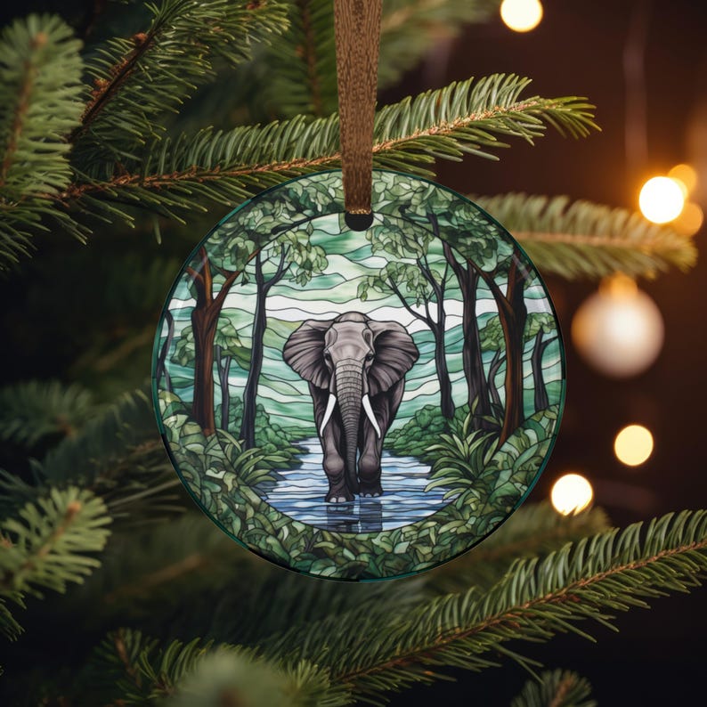 May include: A round glass ornament featuring an elephant walking in a river surrounded by trees. The elephant is gray with white tusks. The background shows a forest scene with green and blue hues. The ornament hangs from a brown ribbon.