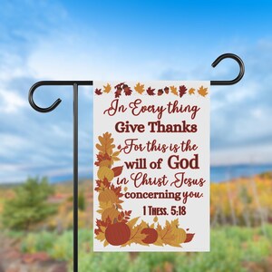 May include: A white garden flag with the text "In Everything Give Thanks" and a Bible verse. The flag features fall-themed graphics of pumpkins and leaves in shades of orange, brown, and yellow. The flag is displayed on a black metal stand.