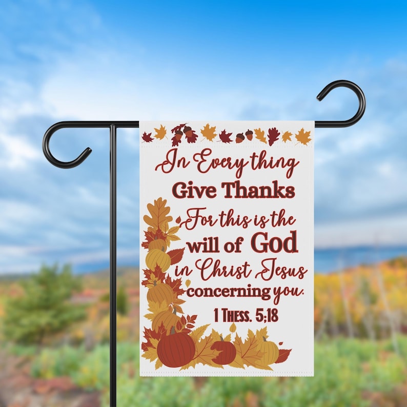 May include: A white garden flag with the text "In Everything Give Thanks" and a Bible verse. The flag features fall-themed graphics of pumpkins and leaves in shades of orange, brown, and yellow. The flag is displayed on a black metal stand.