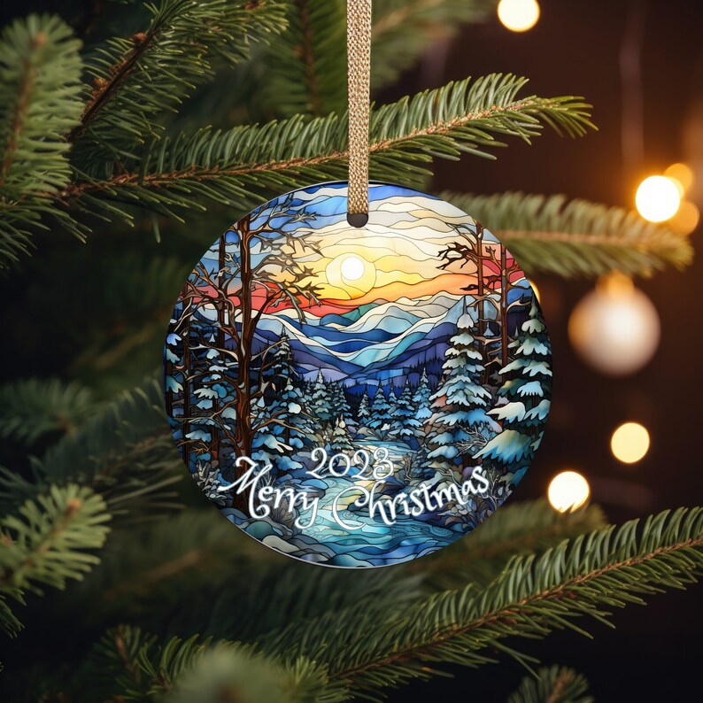 May include: A round Christmas ornament with a stained glass design of a winter mountain scene. The ornament features a blue, green, and white color scheme with a sunset in the background. The text "2023 Merry Christmas" is written in white on the ornament.