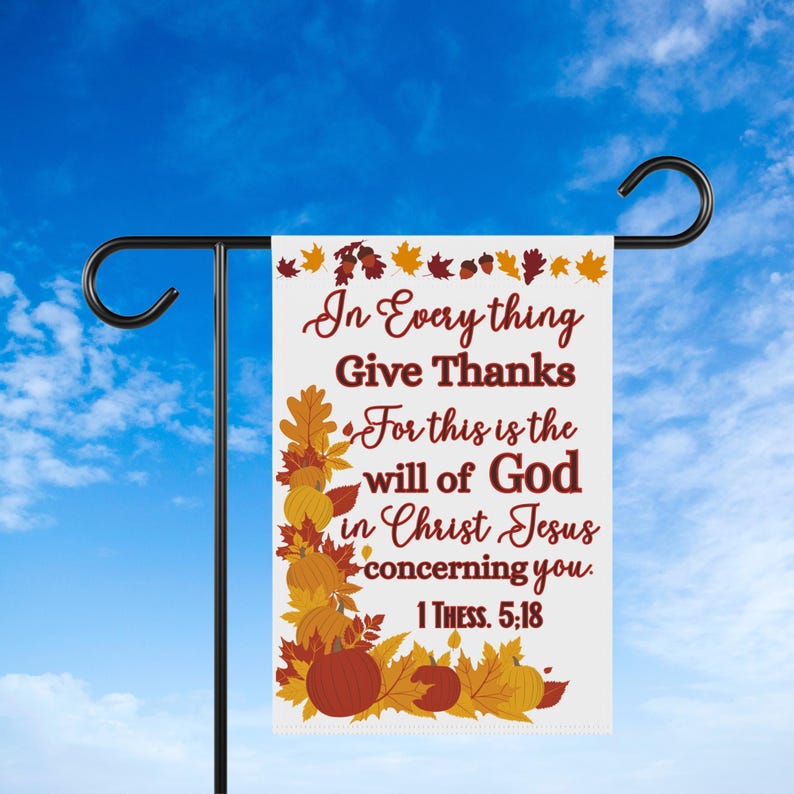 May include: A white garden flag with the text "In Everything Give Thanks" in burgundy script. The flag is decorated with fall leaves and pumpkins in shades of orange, yellow, and brown. The flag is hanging from a black metal garden stake.