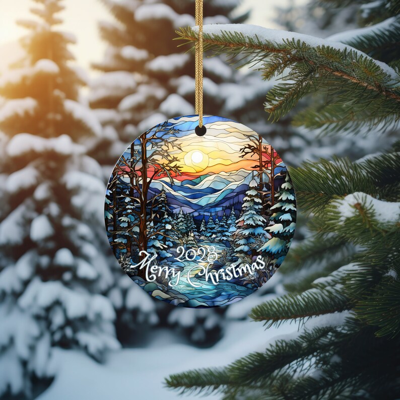 May include: A round Christmas ornament with a stained glass design of a winter forest scene. The ornament features a blue river, snow-covered trees, and a sunset in the background. The text "2023 Merry Christmas" is written on the ornament.