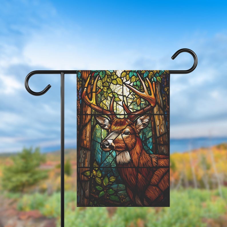 May include: A garden flag displaying a stained-glass style image of a deer with large antlers. The deer is set against a backdrop of a forest with trees and foliage in shades of brown, green, and blue. The flag is mounted on a black metal stand.