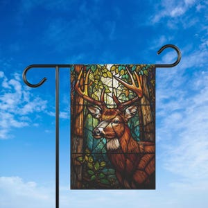 May include: A garden flag with a stained-glass effect depicting a deer with antlers in a forest. The flag is mounted on a black metal pole and hook, set against a blue sky with clouds.