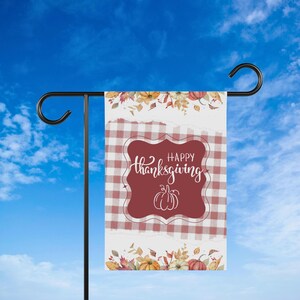 K&ouml;nnte beinhalten: Eine rechteckige Gartenflagge mit einem rot-wei&szlig; karierten Muster und einem weinroten zentralen Design. Das Design zeigt die Worte "Happy Thanksgiving" in Schreibschrift und eine K&uuml;rbisillustration. Florale und K&uuml;rbisakzente zieren die oberen und unteren R&auml;nder.
