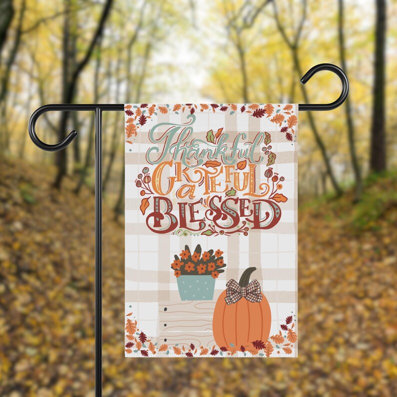May include: A decorative garden flag with the words "Thankful, Grateful, Blessed" in a fall-themed design. The flag features a plaid background, a pumpkin with a bow, and a flower pot. The flag is displayed on a black metal stand.