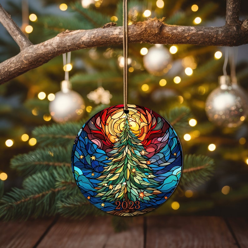 Puede incluir: Un adorno navide&ntilde;o redondo con un dise&ntilde;o de vitral de un &aacute;rbol de Navidad verde con un sol amarillo y un cielo azul. El adorno tiene el a&ntilde;o 2023 escrito en &eacute;l.