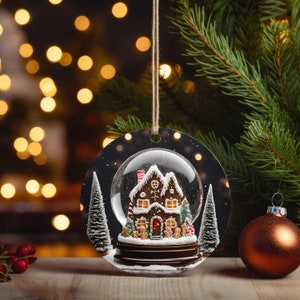 K&ouml;nnte beinhalten: Runder Weihnachtsschmuck mit einem Lebkuchenhaus in einer Schneekugel. Das Haus hat wei&szlig;e Glasur, rote Akzente und ist von kleinen Lebkuchenm&auml;nnchen und schneebedeckten B&auml;umen umgeben. Der Schmuck h&auml;ngt an einer Schnur.