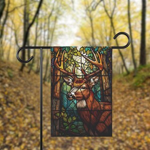 May include: A garden flag with a stained-glass effect depicting a deer with large antlers. The deer is set in a forest scene with trees and foliage in brown, green, and blue hues. The flag is displayed on a black metal stand, suitable for outdoor use.