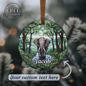 May include: Round Christmas ornament featuring an elephant standing in a river within a forest scene. The ornament has a brown ribbon for hanging and includes the text "Jacob" and "Your custom text here". The ornament is in a free gift box.