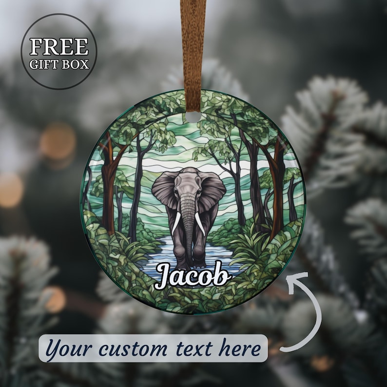 May include: Round Christmas ornament featuring an elephant standing in a river within a forest scene. The ornament has a brown ribbon for hanging and includes the text "Jacob" and "Your custom text here". The ornament is in a free gift box.