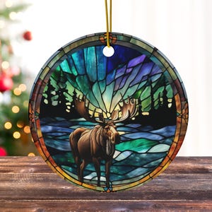 K&ouml;nnte beinhalten: Ein Weihnachtsbaumschmuck im Stil eines Buntglasfensters mit einem Elch, der in einem schneebedeckten Wald steht, vor dem Hintergrund einer farbenfrohen Aurora Borealis.