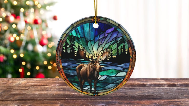 K&ouml;nnte beinhalten: Ein Weihnachtsbaumschmuck im Stil eines Buntglasfensters mit einem Elch, der in einem schneebedeckten Wald steht, vor dem Hintergrund einer farbenfrohen Aurora Borealis.
