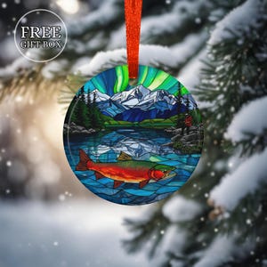 May include: Round Christmas ornament with a nature scene featuring a lake, mountains, and a colorful fish. The ornament has a red ribbon for hanging and the text "FREE GIFT BOX" in a white circle.