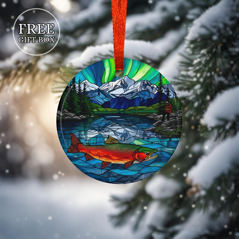 May include: Round Christmas ornament with a nature scene featuring a lake, mountains, and a colorful fish. The ornament has a red ribbon for hanging and the text "FREE GIFT BOX" in a white circle.