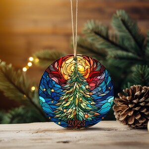 Puede incluir: Un adorno navide&ntilde;o redondo con un dise&ntilde;o de vitral de un &aacute;rbol de Navidad con un sol y estrellas. El adorno es azul, verde, amarillo, rojo y naranja. El a&ntilde;o 2023 est&aacute; escrito en la parte inferior del adorno.