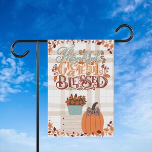 May include: A decorative garden flag with the words "Thankful, Grateful, Blessed" in a fall-themed design. The flag features a pumpkin, flowers, and autumn leaves against a plaid background. The flag is displayed on a black metal stand.