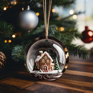 K&ouml;nnte beinhalten: Eine transparente, runde Weihnachtskugel mit einem Lebkuchenhaus im Inneren, komplett mit wei&szlig;em Dach, roter T&uuml;r und gr&uuml;nen B&auml;umen. Der Schmuck tr&auml;gt die Aufschrift "Merry Christmas" und h&auml;ngt an einer braunen Schnur.