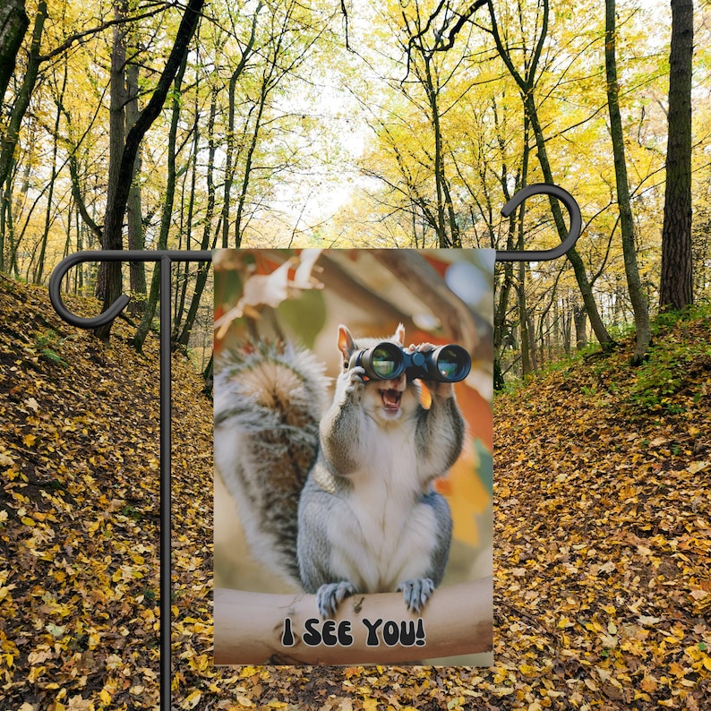 May include: A garden flag with a squirrel wearing binoculars and the text "I see you!" The squirrel is sitting on a branch with a blurred background of fall foliage.