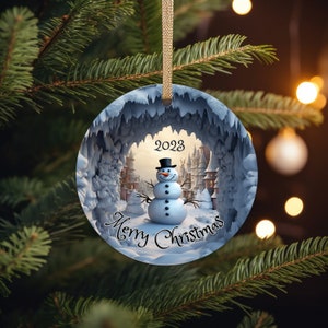 Puede incluir: Un adorno navide&ntilde;o redondo con un mu&ntilde;eco de nieve de pie en una escena nevada. El mu&ntilde;eco de nieve lleva un sombrero de copa negro y tiene una nariz de bot&oacute;n negro. El adorno tiene el texto "2023 Feliz Navidad" en &eacute;l.