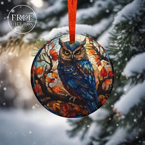 May include: Round Christmas ornament featuring a blue owl perched on a branch with autumn leaves. The ornament has a stained-glass effect and is suspended by a red ribbon. The text "FREE GIFT BOX" is in the upper left corner.