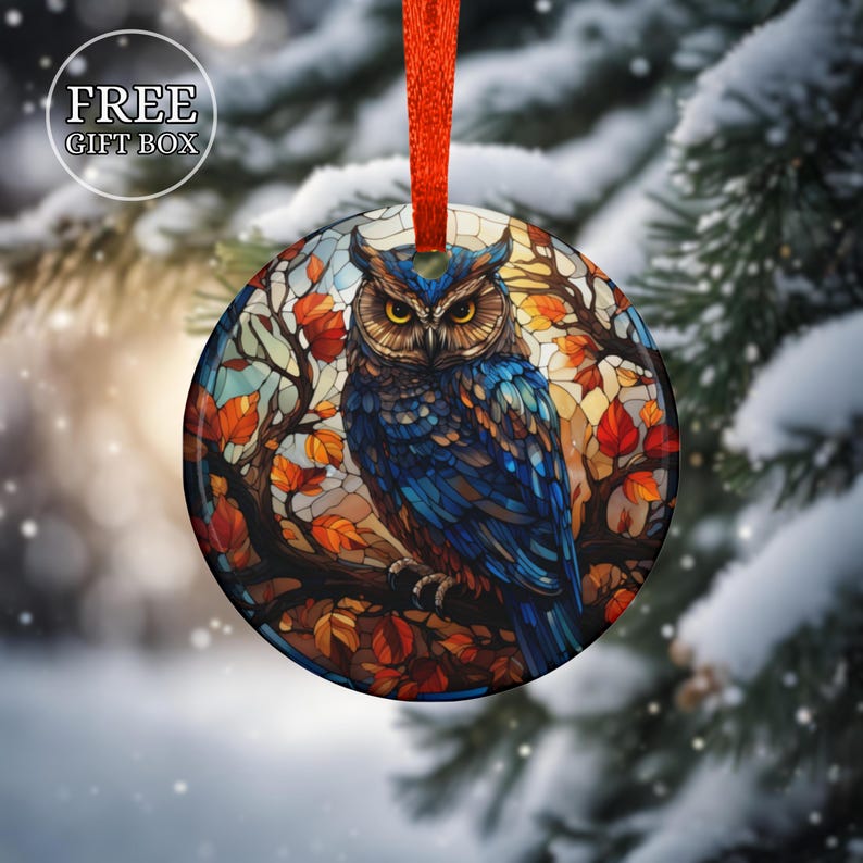 May include: Round Christmas ornament featuring a blue owl perched on a branch with autumn leaves. The ornament has a stained-glass effect and is suspended by a red ribbon. The text "FREE GIFT BOX" is in the upper left corner.