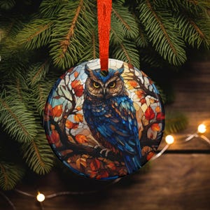 May include: Round Christmas ornament featuring a blue owl perched on a branch with autumn leaves. The owl has yellow eyes and is set against a stained-glass style background. The ornament is hanging from a red ribbon.