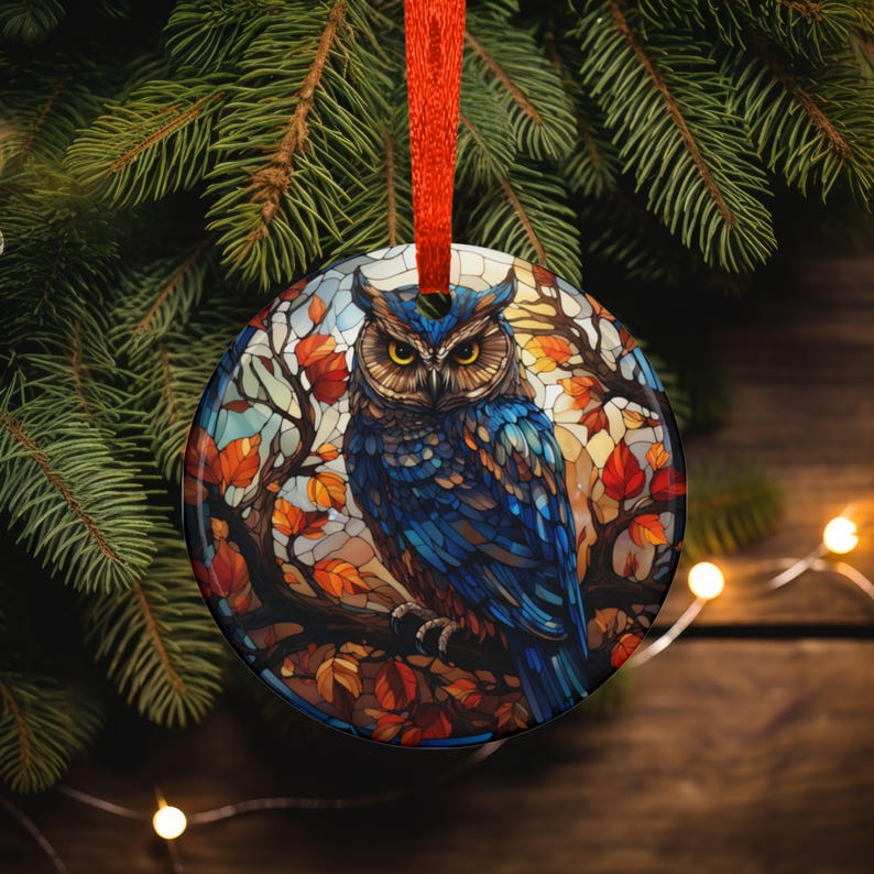 May include: Round Christmas ornament featuring a blue owl perched on a branch with autumn leaves. The owl has yellow eyes and is set against a stained-glass style background. The ornament is hanging from a red ribbon.