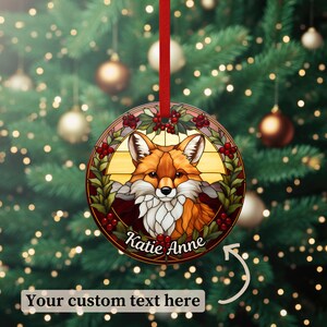 May include: A round glass ornament with a stained glass design of a fox in a wreath of holly and berries. The ornament is red, green, and gold and has the text "Katie Anne" at the bottom.