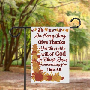 May include: A white garden flag with the text "In Everything Give Thanks" in burgundy script. The flag features autumn leaves and pumpkins in shades of orange, brown, and yellow. The flag is displayed on a black metal stand.