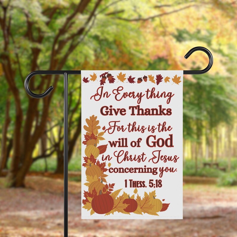 May include: A white garden flag with the text "In Everything Give Thanks" in burgundy script. The flag features autumn leaves and pumpkins in shades of orange, brown, and yellow. The flag is displayed on a black metal stand.