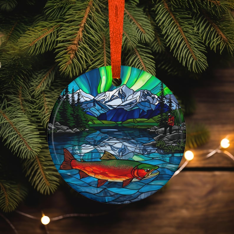 May include: Round Christmas ornament with a stained-glass style design. The ornament features a mountain lake scene with a red and green fish, a person fishing, and the Northern Lights. The ornament is hanging from an orange ribbon.
