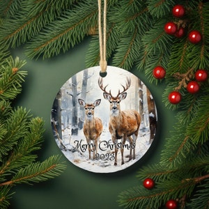 May include: Round Christmas ornament featuring a winter scene with two deer in a snowy forest. The ornament is white with a brown border and the text "Merry Christmas 2023" printed on the bottom.