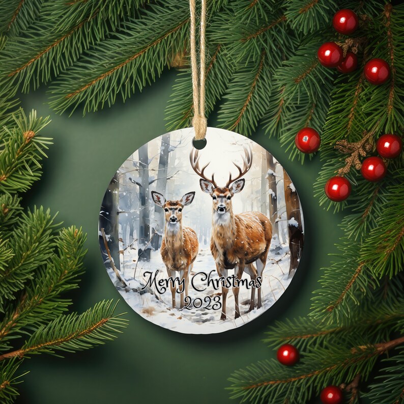 May include: Round Christmas ornament featuring a winter scene with two deer in a snowy forest. The ornament is white with a brown border and the text "Merry Christmas 2023" printed on the bottom.