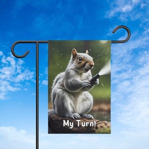 May include: A gray squirrel holds a garden hose and looks at the camera. The squirrel is sitting on a rock with green foliage behind it. The text "My Turn!" is printed on the rock.